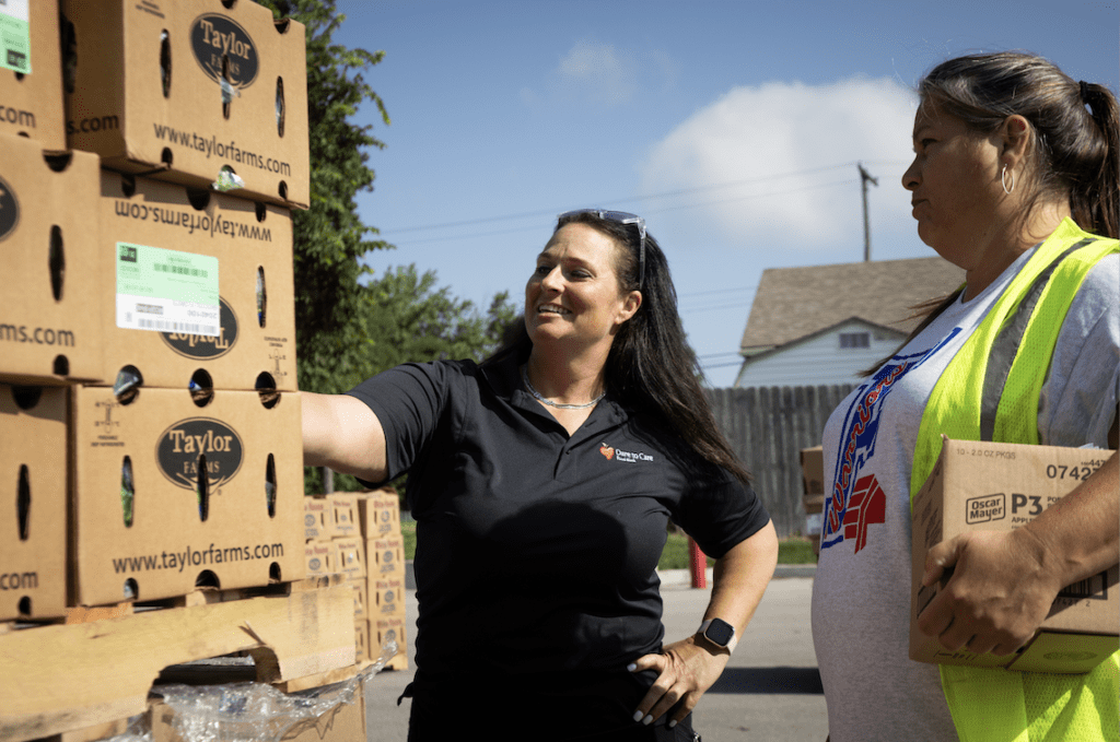 A person in a black polo shirt is smiling and pointing at a sign on a wooden pole next to another individual wearing a medal.