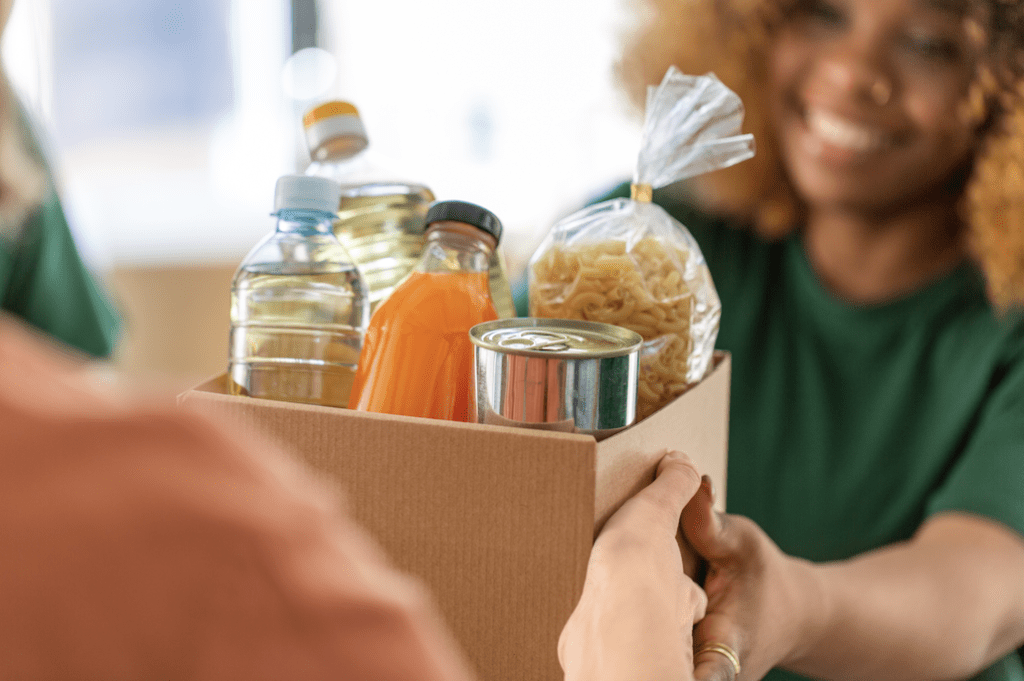 A cardboard box filled with various food items such as water bottles, pasta, and a tin can is being handed to someone.