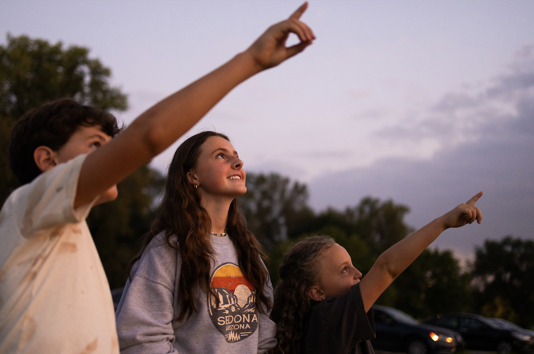 Individuals are looking up towards the sky, with one person pointing upwards, against a backdrop of a dusk sky.