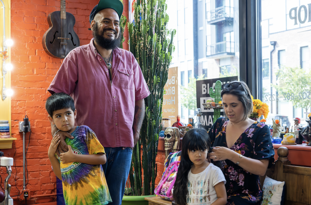 A smiling individual stands in a brightly lit indoor space with plants and a guitar in the background, while others appear engaged in a casual activity nearby.