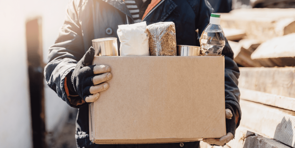 A person is holding a cardboard box containing various food items.