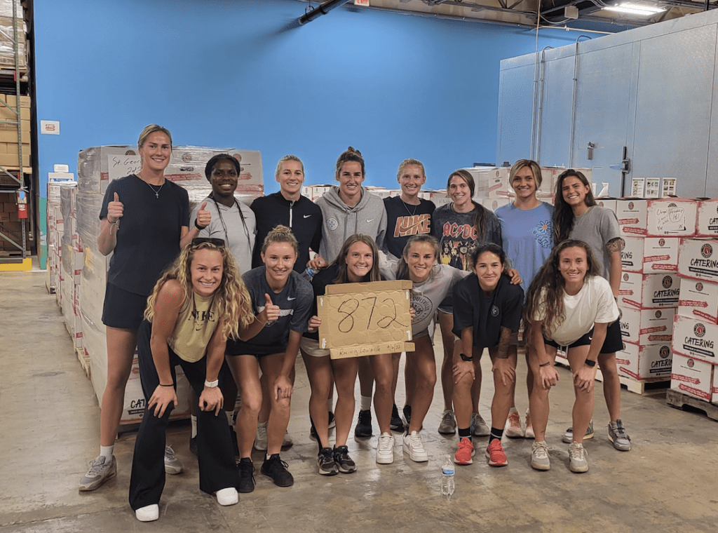 A group of individuals is posing for a photo with some standing and others kneeling, while one person holds a sign displaying 