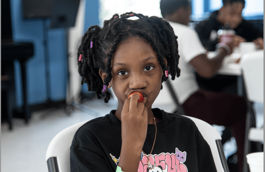 A young individual with braided hair and colorful ties is sitting and biting into a red fruit while looking thoughtfully to the side.