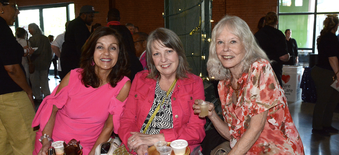 Three individuals are seated side by side, holding drinks and smiling for the camera at an event.