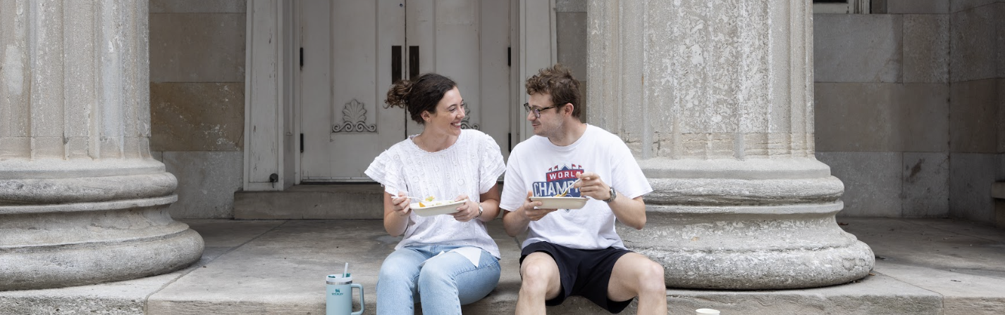 Two individuals sit on outdoor steps, enjoying a meal and smiling at each other.